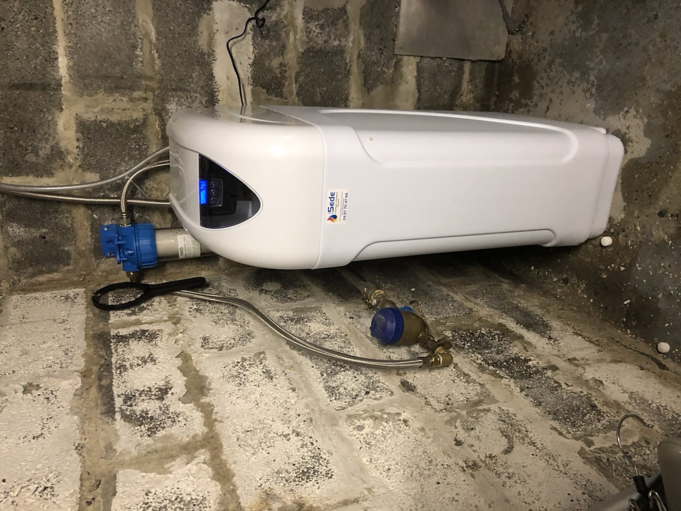 Un adoucisseur d'eau blanc avec écran bleu est posé dans un local en parpaings, à côté d'un filtre bleu et de flexibles métalliques posés au sol.