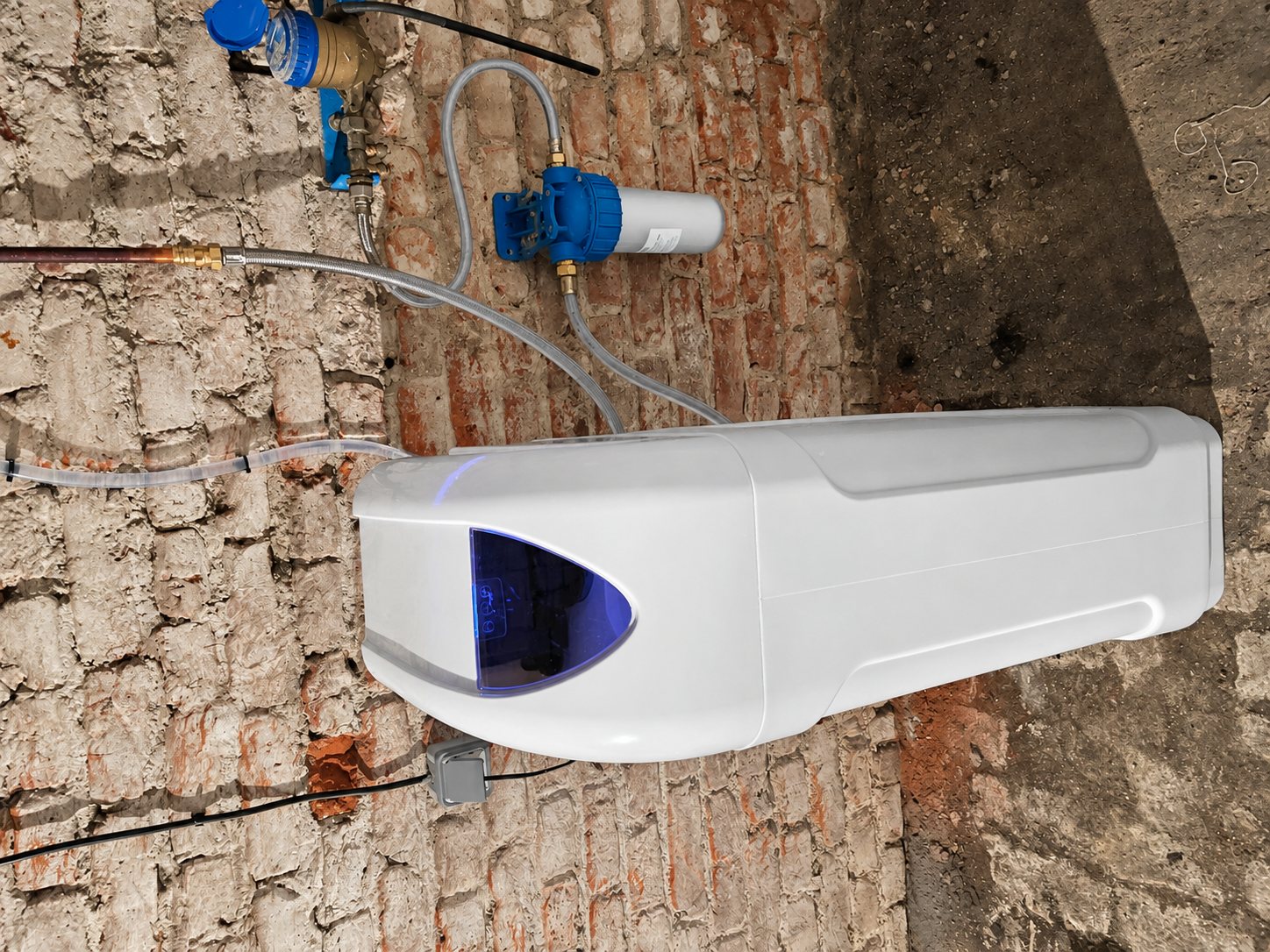 Un adoucisseur d'eau blanc à écran bleu lumineux est installé contre un mur de briques anciennes, relié à des tuyaux et à un filtre bleu.