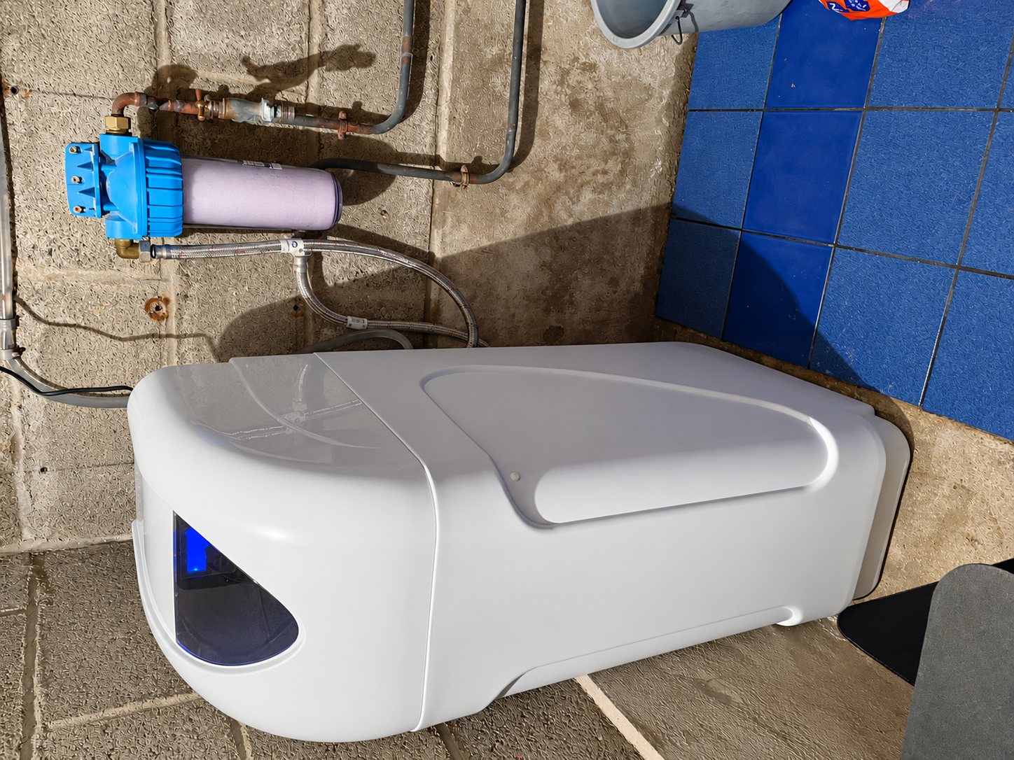 Un adoucisseur d'eau blanc posé au sol dans un local technique en parpaings, à côté d'un filtre à cartouche à carter bleu raccordé par des tuyaux.