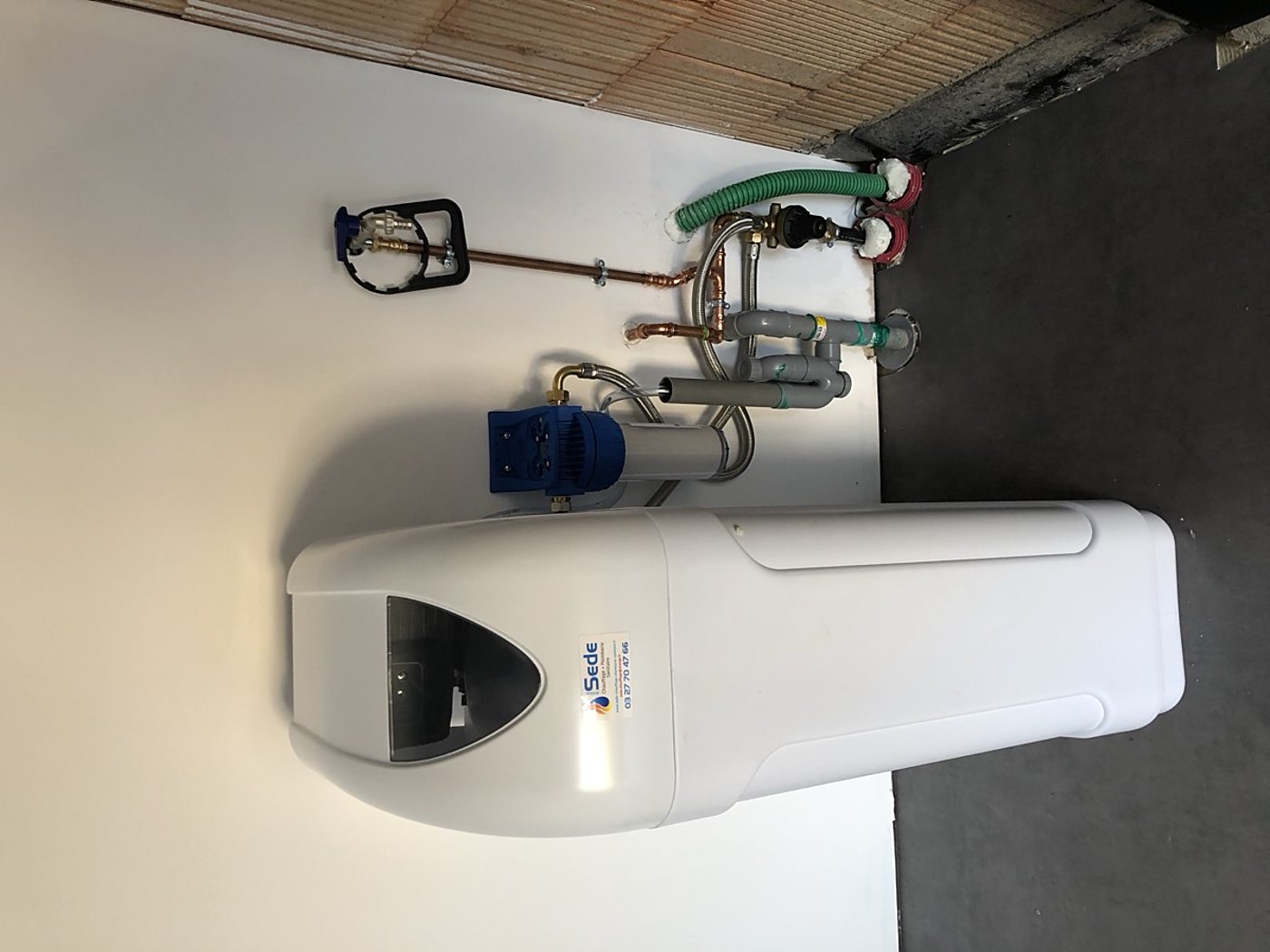 Un adoucisseur d'eau blanc de la marque Sede, posé au sol contre un mur blanc et raccordé à des tuyaux en cuivre et en PVC gris.