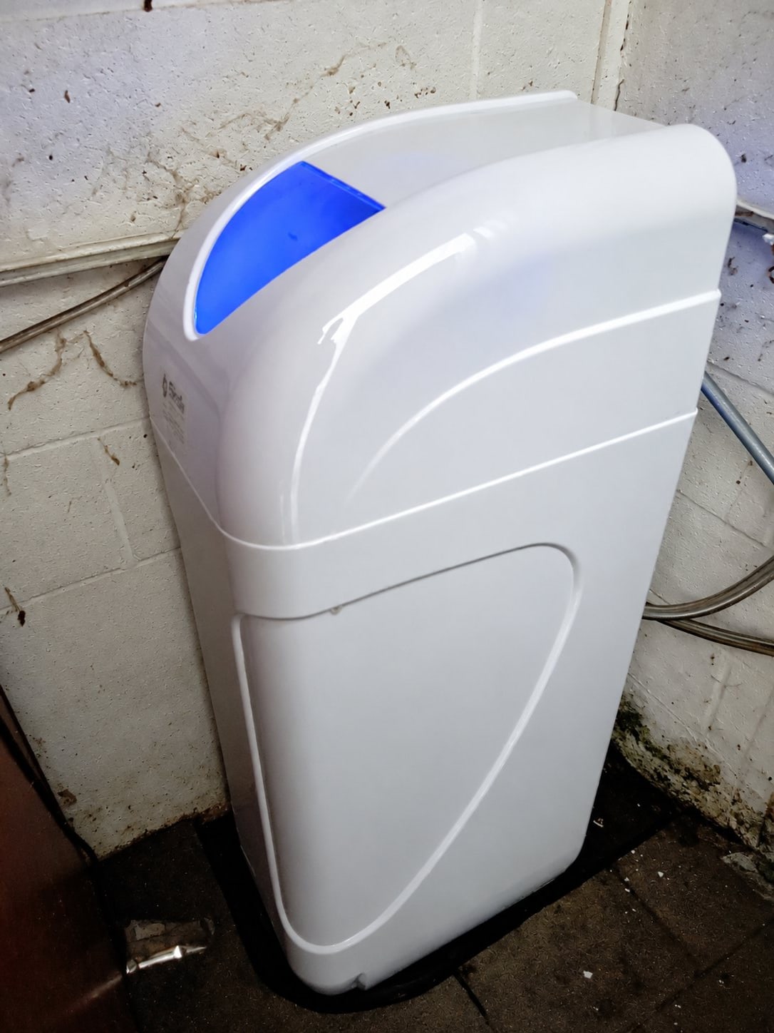 Un boîtier blanc d'adoucisseur d'eau avec un voyant bleu allumé est posé sur un sol carrelé près de câbles et de tuyaux.