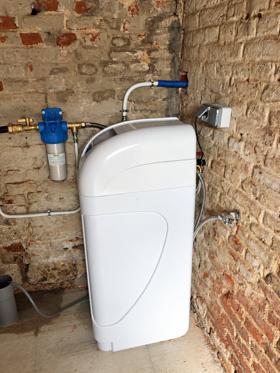 Un adoucisseur d'eau blanc est fixé dans une cave aux murs de briques apparentes, relié par des tuyaux à un filtre bleu et à une prise électrique.