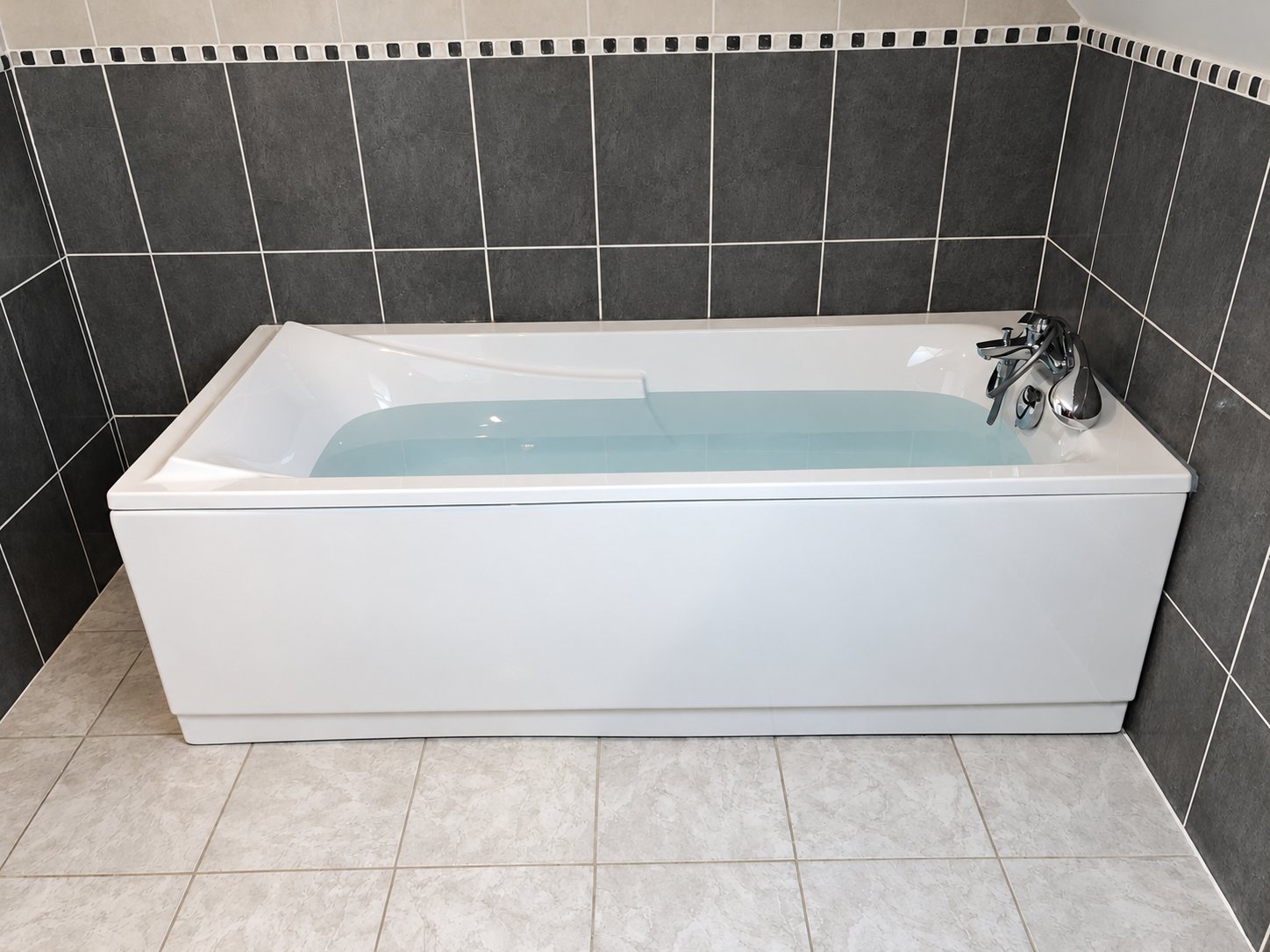Une baignoire rectangulaire blanche remplie d'eau, équipée d'un mitigeur chromé, installée contre un mur de carreaux gris foncé avec frise de mosaïque.