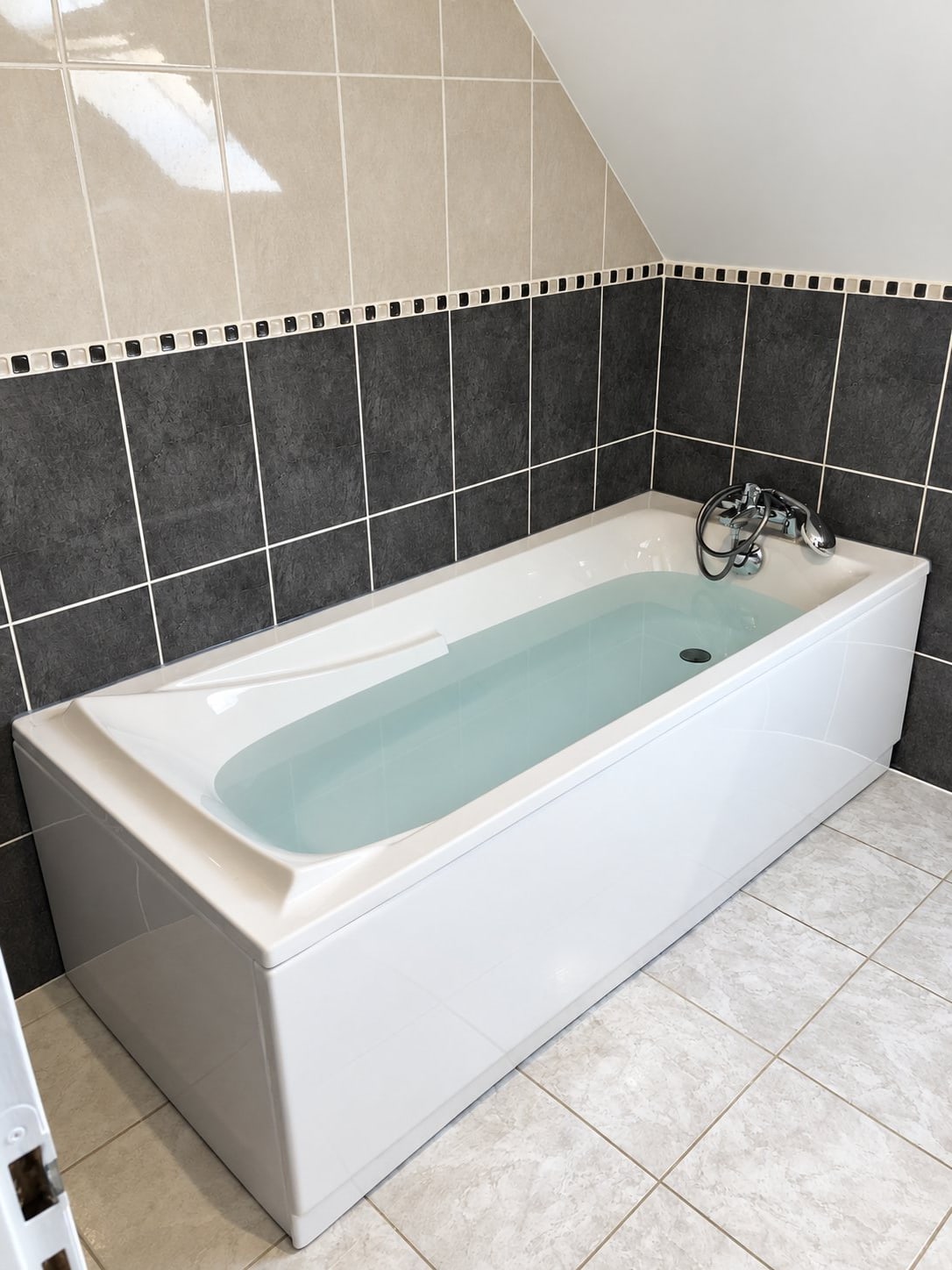 Une baignoire blanche avec mitigeur chromé est installée dans une salle de bains rénovée, entourée de carreaux gris foncé, beiges et gris clair avec une frise de mosaïque.