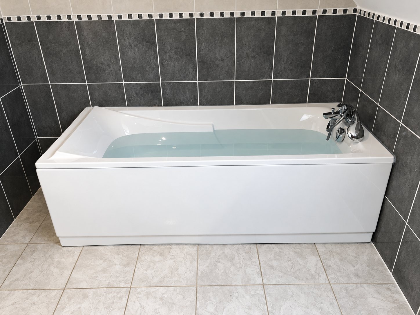 Une baignoire rectangulaire blanche remplie d'eau, équipée d'une robinetterie chromée, est encastrée dans une salle de bains aux murs de carreaux gris foncé et au sol de carreaux gris clair.