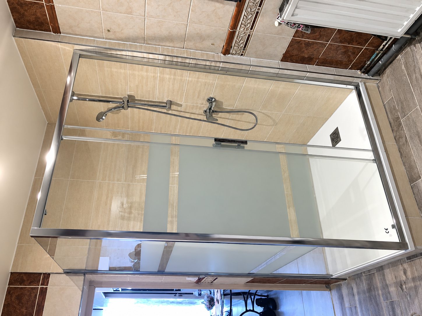 Une cabine de douche en verre partiellement dépoli, avec cadre chromé et barre de douche, installée dans une salle de bain carrelée beige et marron.