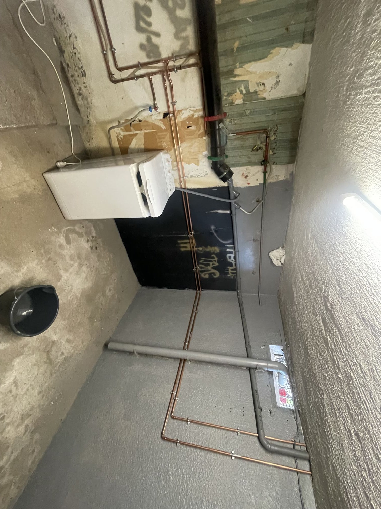 Dans une cave aux murs gris et noirs, un lave-linge blanc est installé près de tuyaux en cuivre et de conduites grises, avec un seau noir au sol.