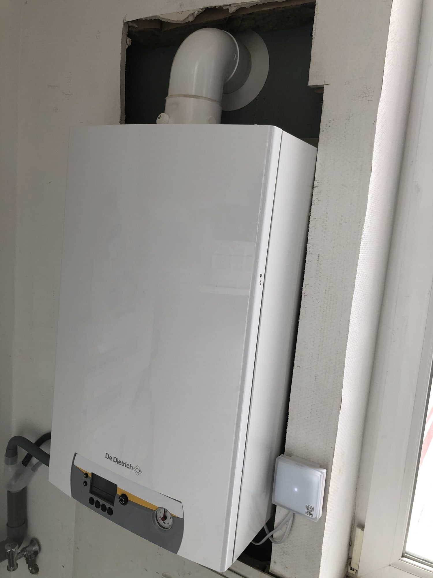 Une chaudière murale blanche De Dietrich est installée sous une fenêtre, raccordée à un conduit d'évacuation blanc et à des tuyaux, avec un petit thermostat sans fil au-dessus.