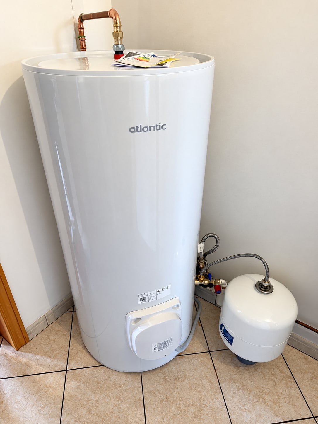 Un chauffe-eau électrique blanc de marque Atlantic, posé sur un sol carrelé et raccordé en cuivre, avec un petit vase d'expansion blanc derrière lui.