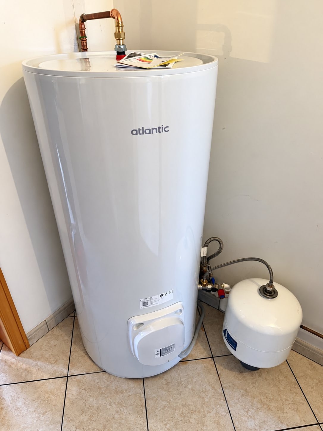Un chauffe-eau électrique blanc de marque Atlantic, raccordé en cuivre, est installé dans un coin avec un vase d'expansion blanc posé derrière lui sur un sol carrelé beige.