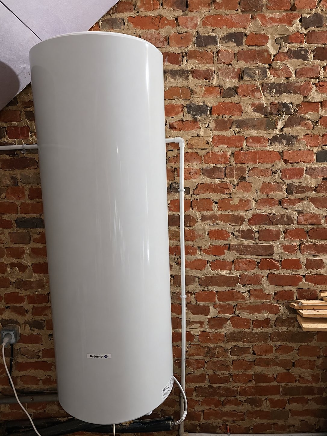 Un grand chauffe-eau électrique blanc de marque De Dietrich est fixé horizontalement devant un mur de briques apparentes, avec un tuyau blanc au-dessus.