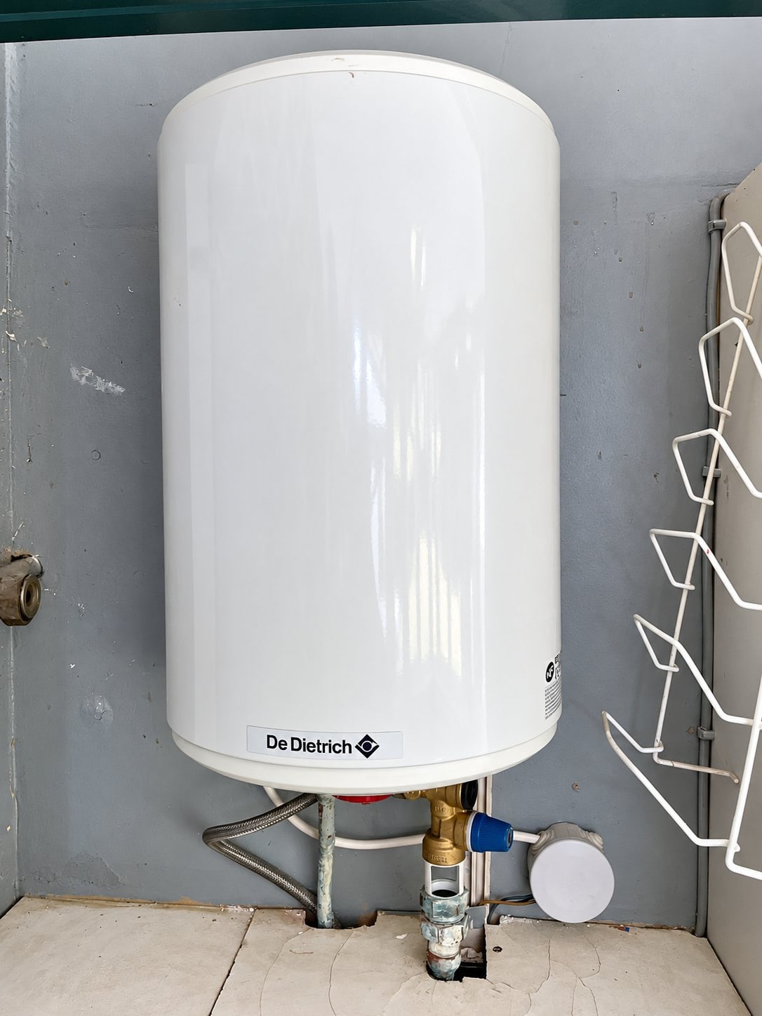 Un chauffe-eau électrique blanc de marque De Dietrich, photographié à l'horizontale, est installé dans un placard avec son groupe de sécurité et ses raccordements d'eau.