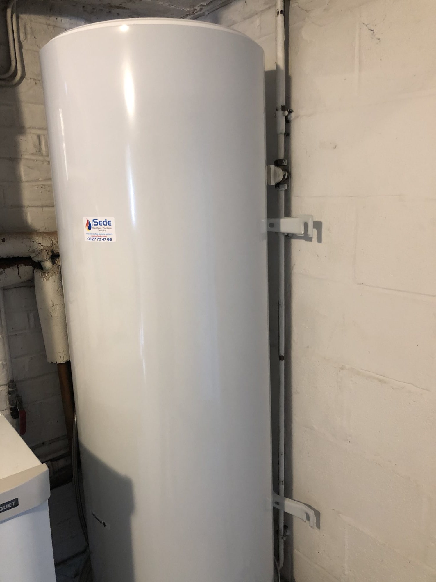 Un grand ballon de chauffe-eau blanc posé à l'horizontale, fixé contre un mur dans un local technique, avec une étiquette Sede sur sa paroi.
