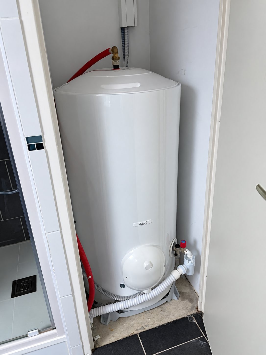 Un ballon d'eau chaude blanc est installé dans un placard technique, raccordé par un flexible rouge et un tuyau d'évacuation blanc.