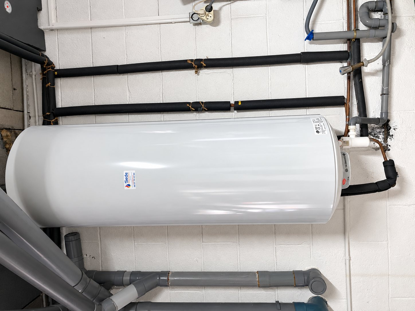 Un ballon d'eau chaude blanc avec une étiquette Sede est installé contre un mur de briques blanches, raccordé à des tuyaux isolés noirs et à des conduits gris.