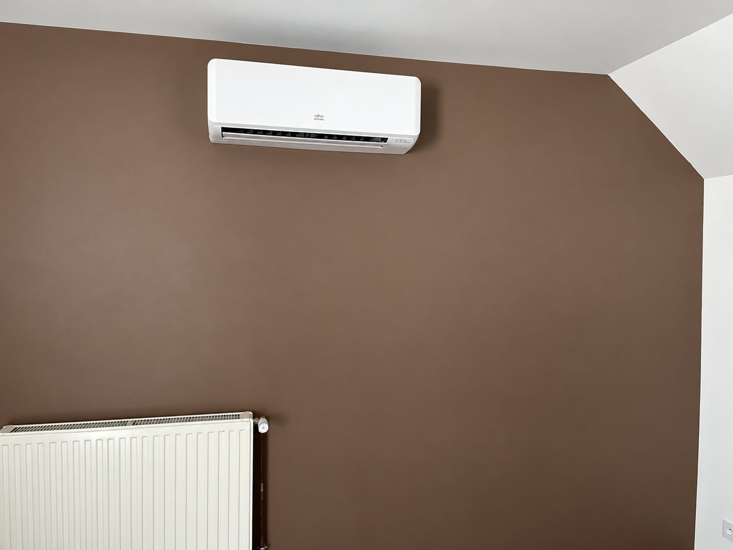 Un climatiseur split blanc fixé en hauteur sur un mur marron, au-dessus d'un radiateur blanc, dans une pièce sous combles.