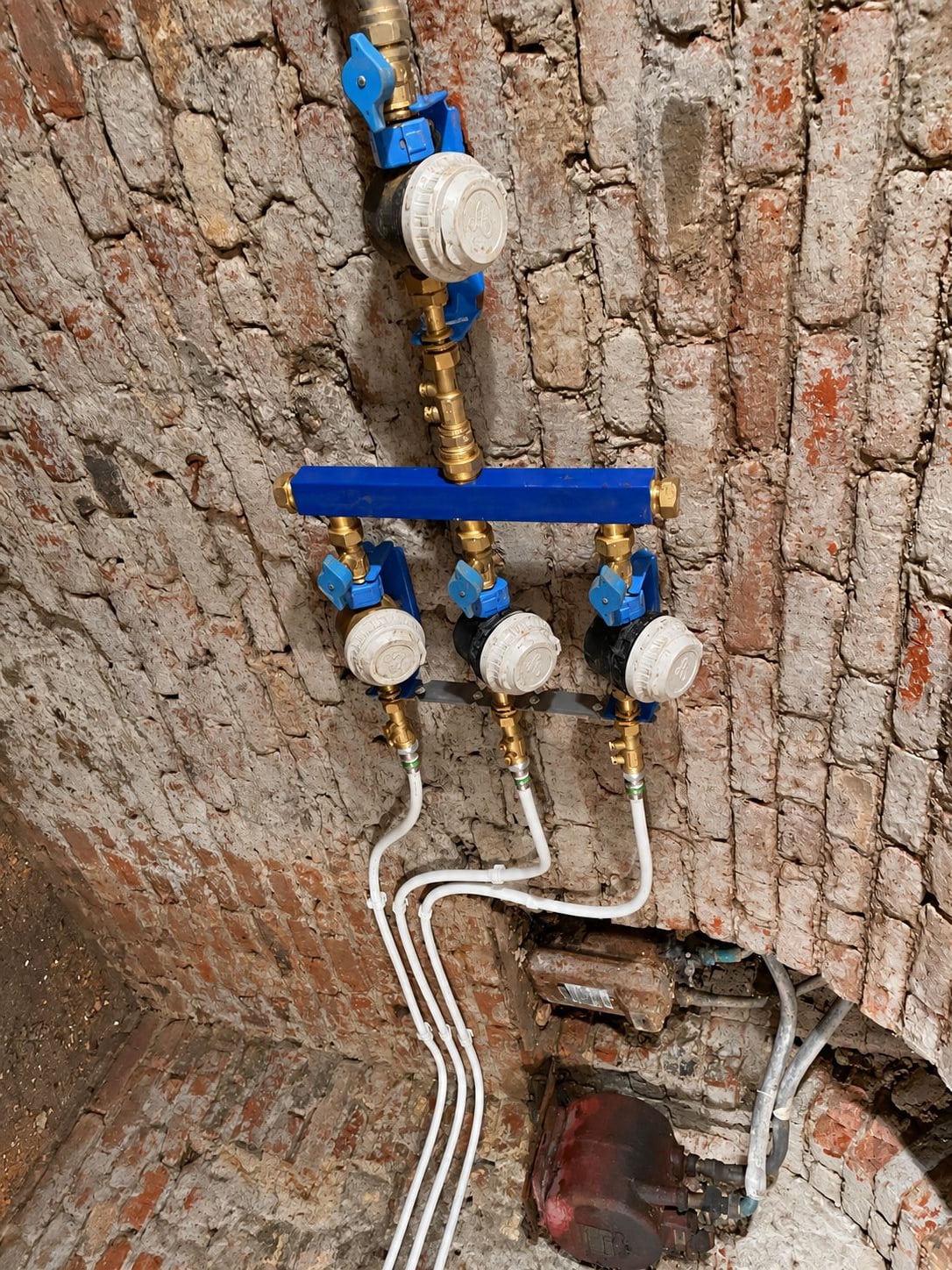 Installation de plomberie dans une cave voûtée en briques, avec un collecteur bleu, quatre compteurs d'eau et des tuyaux blancs.