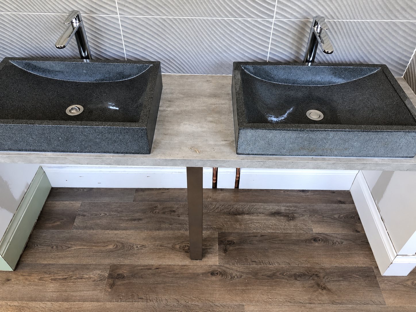 Deux vasques rectangulaires en pierre noire avec robinets chromés posées sur un plan gris, dans une salle de bain au carrelage mural en relief et au sol en parquet stratifié.