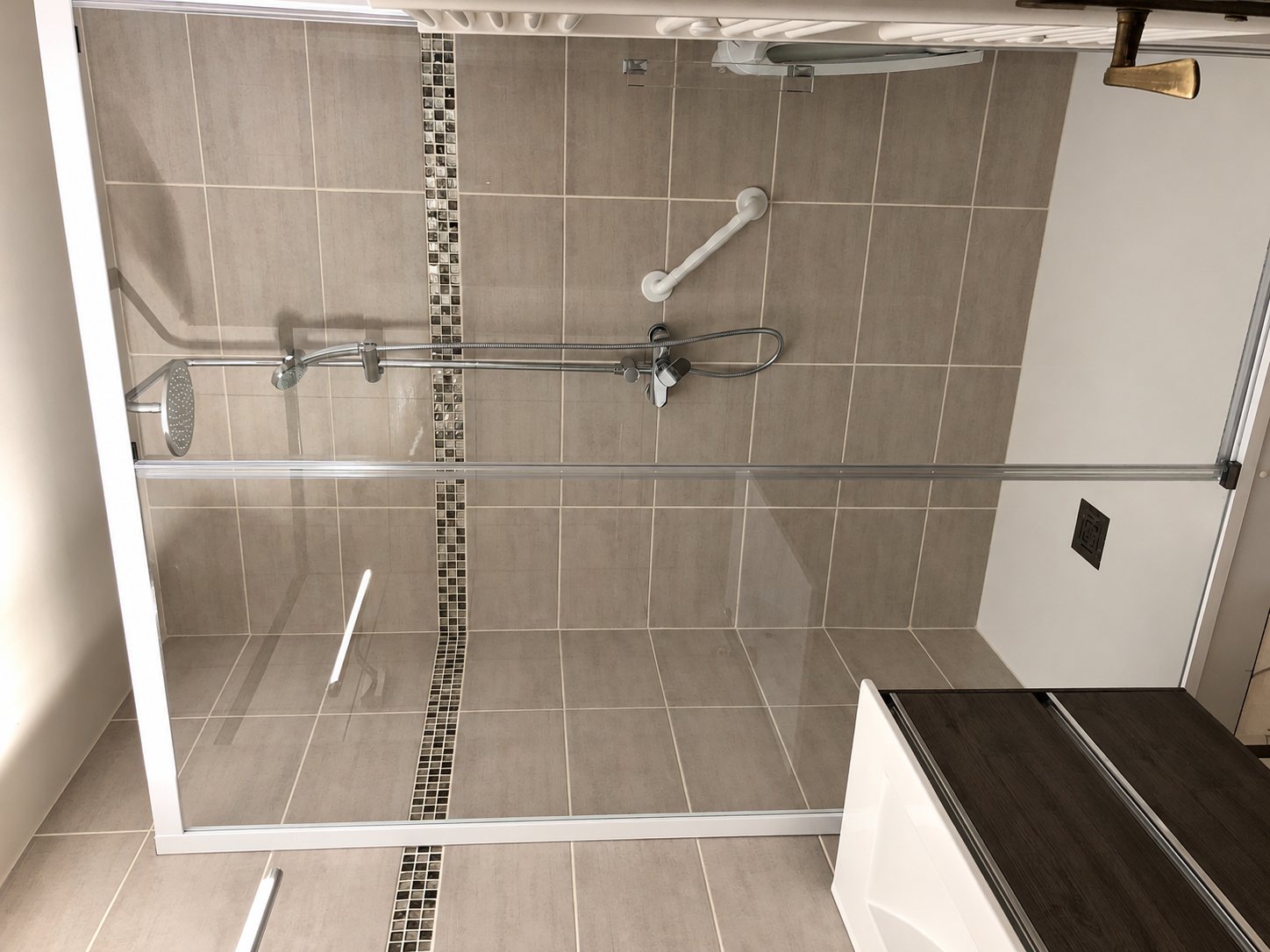 Douche rénovée avec paroi vitrée coulissante, carrelage gris, frise en mosaïque, barre d'appui et pomme de douche ronde.