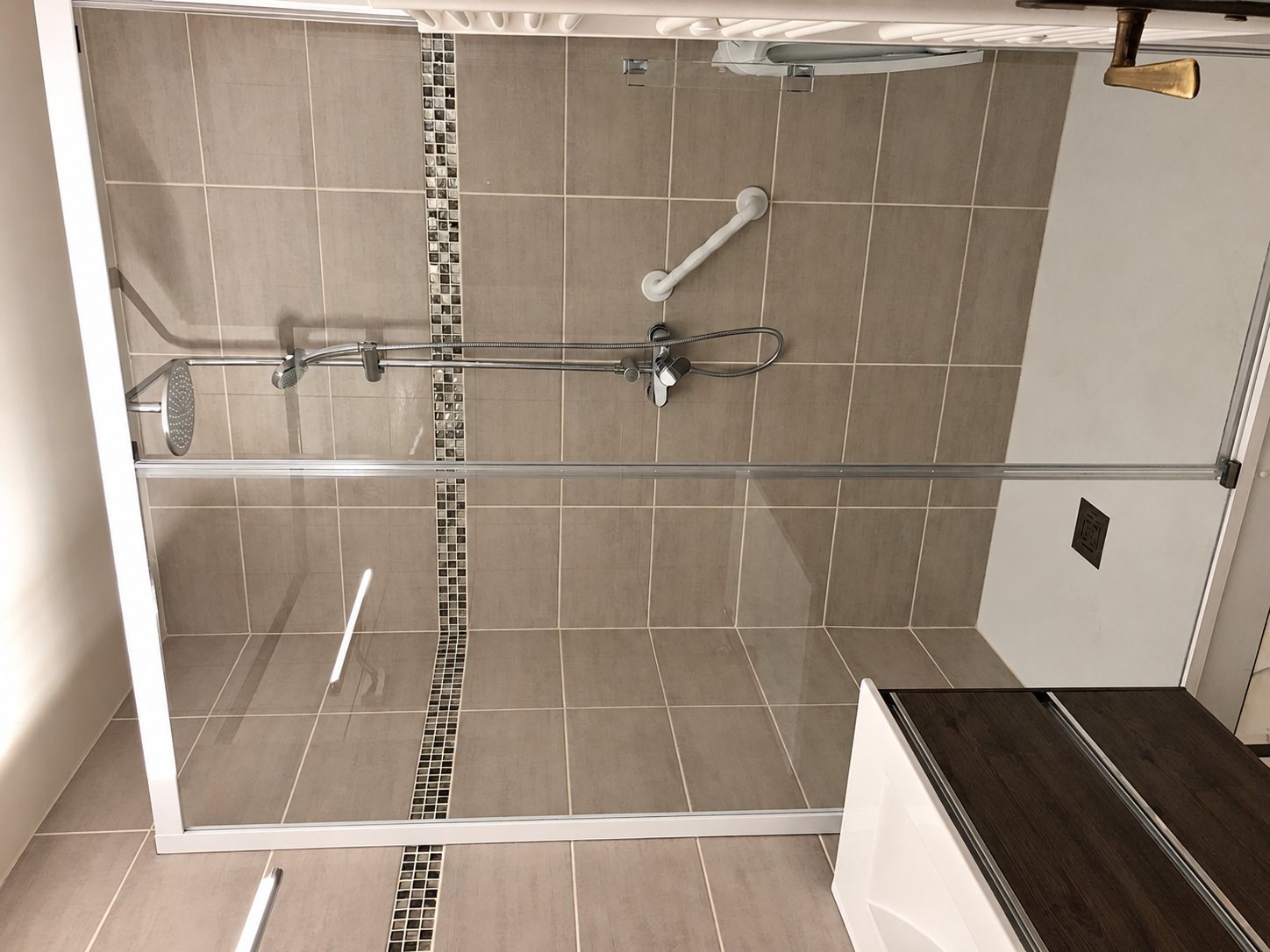 Une douche rénovée avec paroi vitrée, carrelage gris, bande décorative en mosaïque, barre d'appui et pomme de douche.