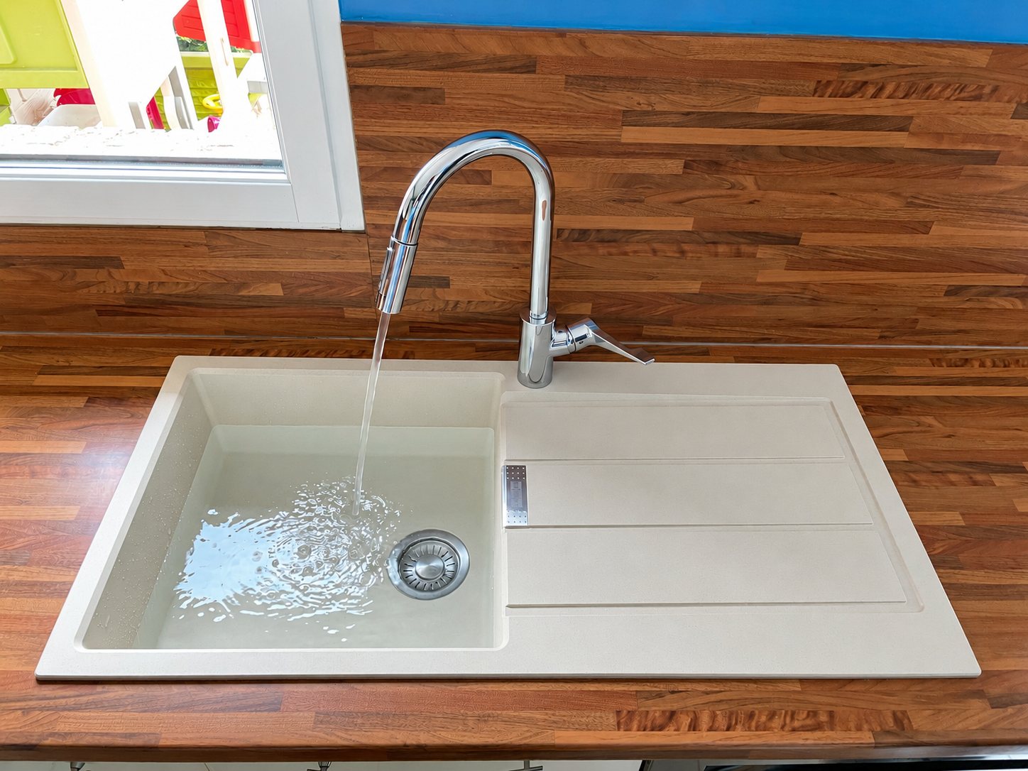 Un évier rectangulaire beige avec égouttoir et robinet chromé à bec haut, dont l'eau coule, encastré dans un plan de travail en bois.