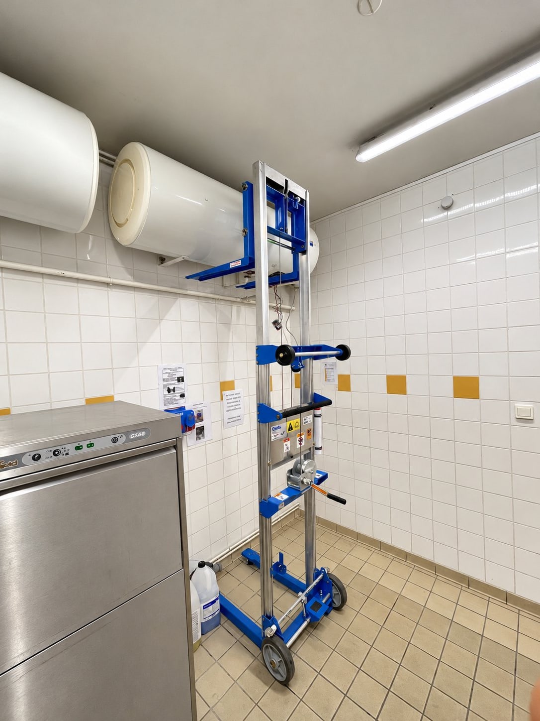 Un monte-matériaux bleu et aluminium sur roues est posé dans un local carrelé de blanc, à côté de deux chauffe-eau et d'un lave-vaisselle professionnel en inox.