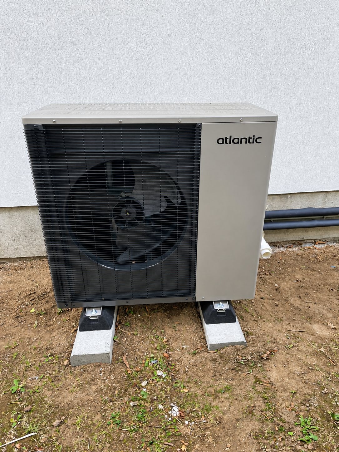 Une pompe à chaleur de marque Atlantic posée sur deux plots en béton contre un mur blanc, sur un sol de terre.