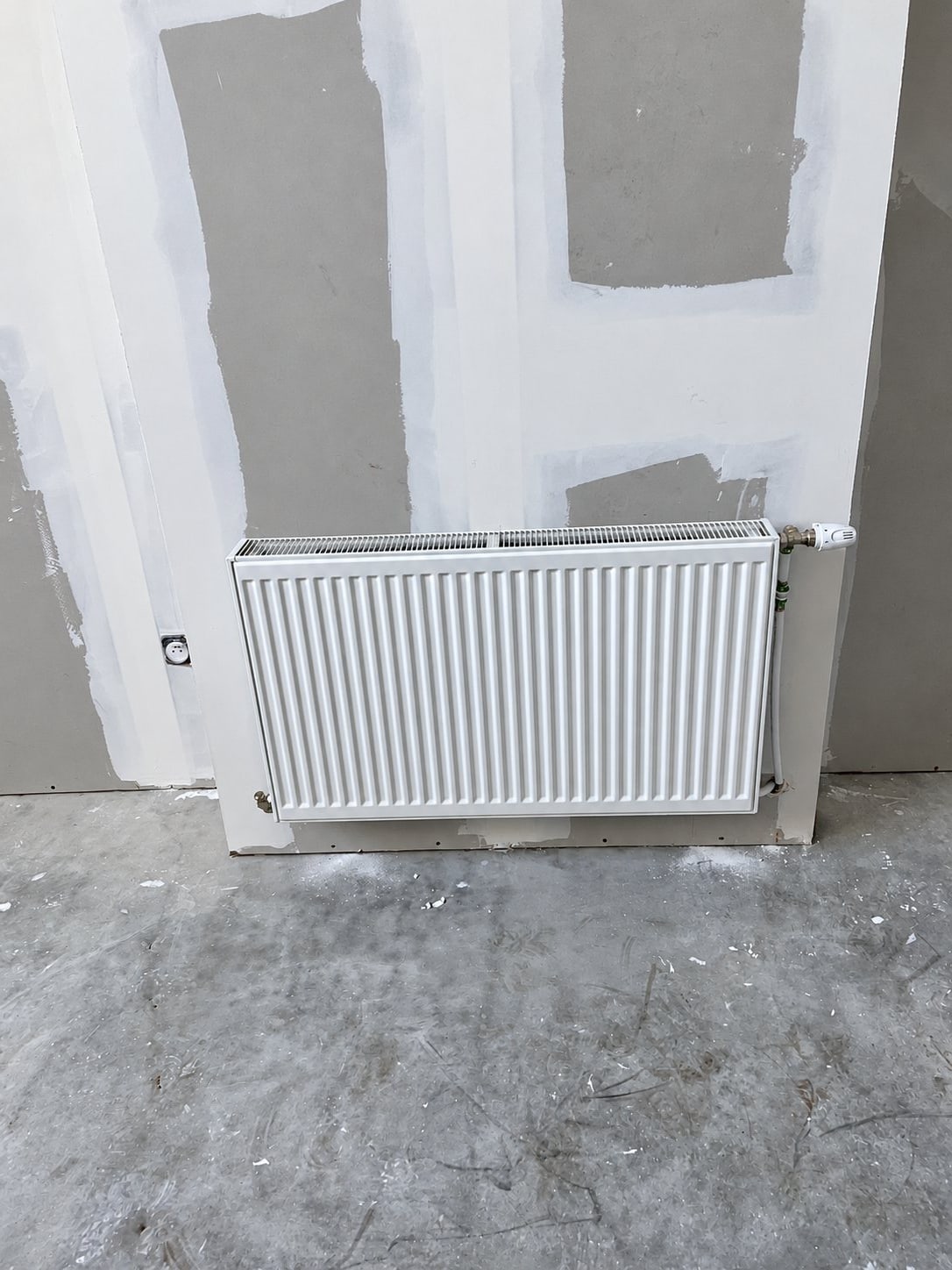 Un radiateur blanc à panneau avec robinet thermostatique est fixé sur un mur en plaques de plâtre enduites, dans un chantier de rénovation au sol en béton gris.