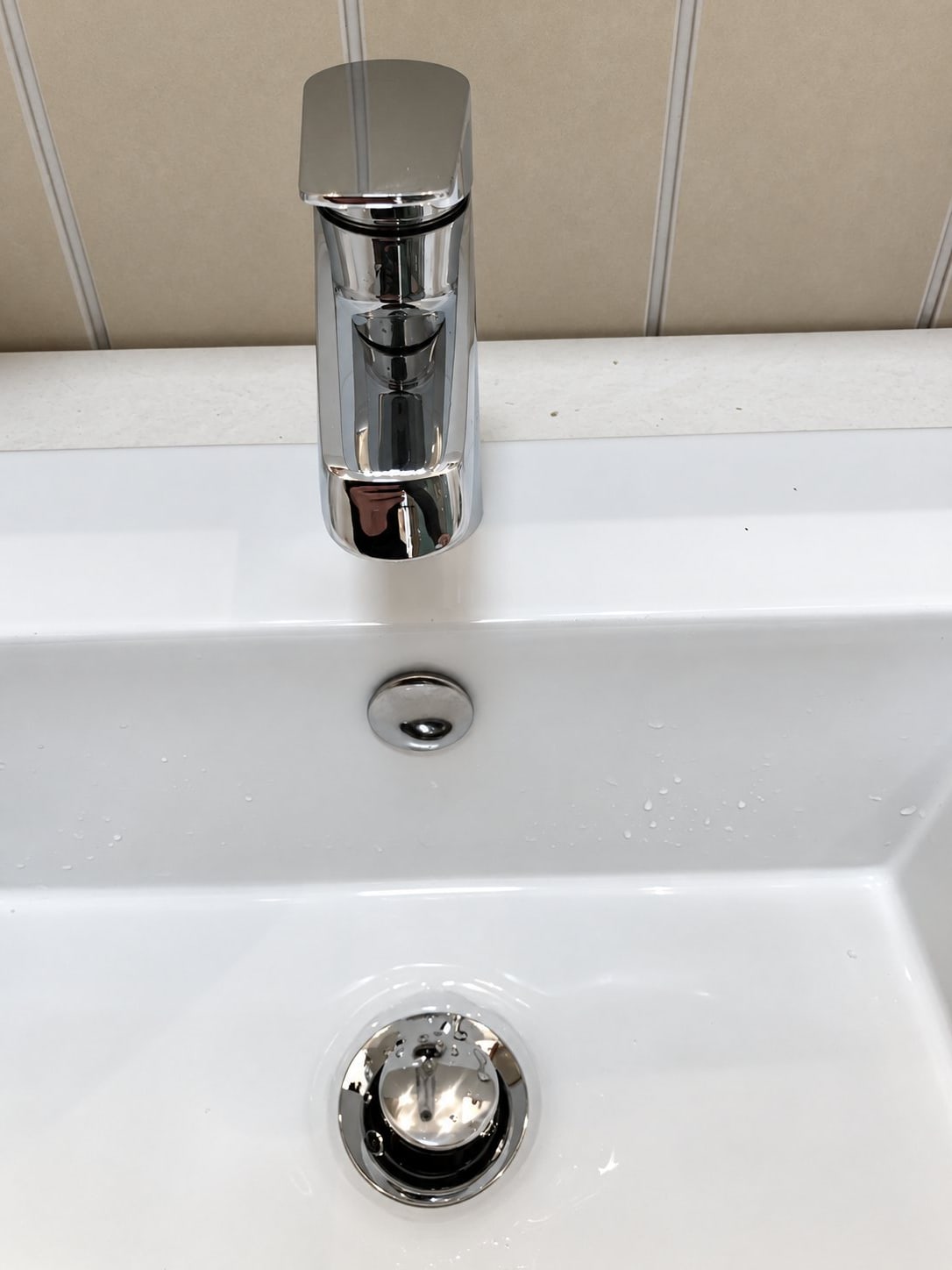 Gros plan sur un robinet chromé moderne posé sur un lavabo blanc, avec un bouton de trop-plein et une bonde chromée, devant un mur lambrissé beige.