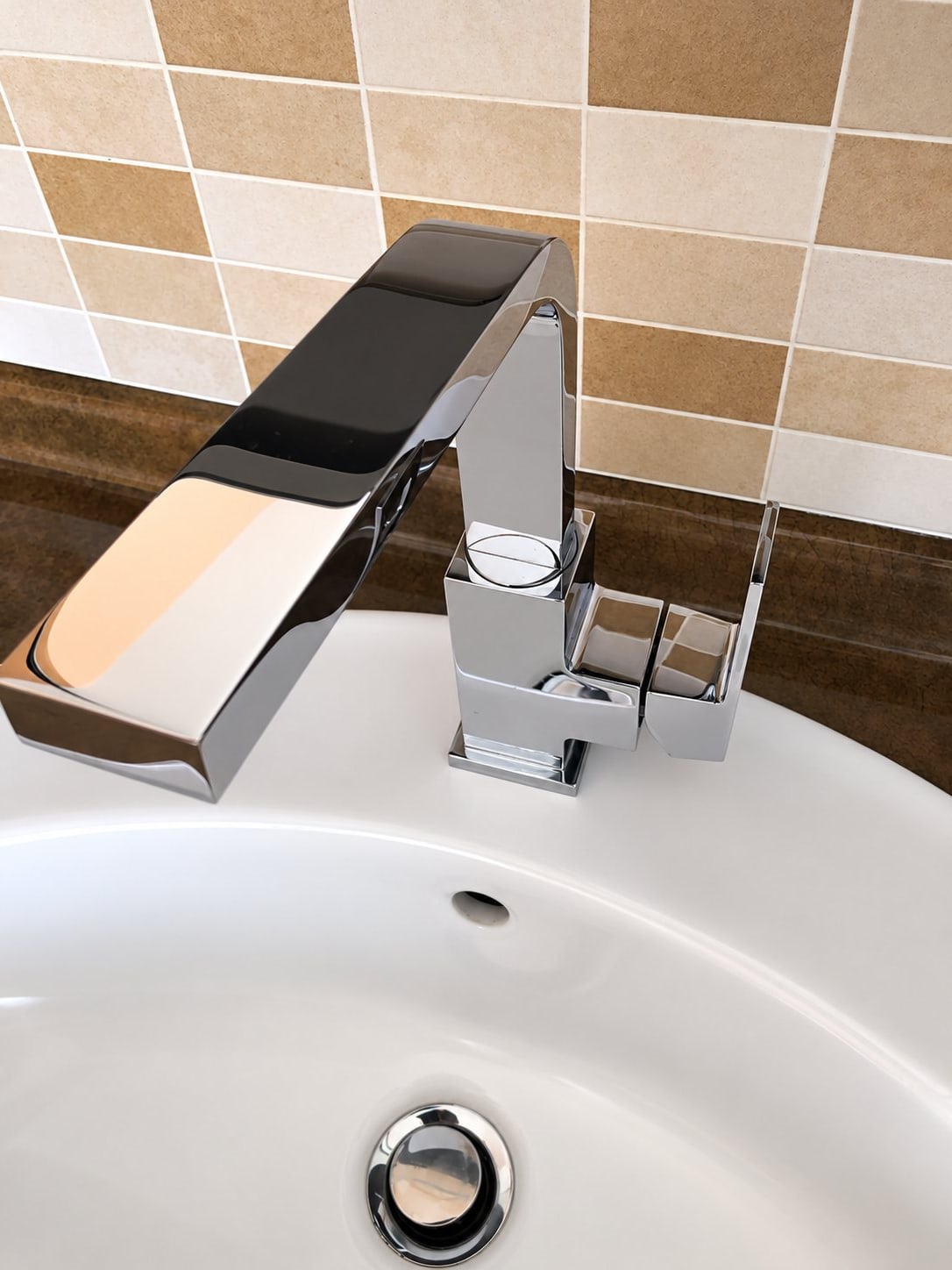 Robinet mitigeur chromé de forme carrée sur un lavabo blanc, devant un mur carrelé de carreaux beiges et marron.