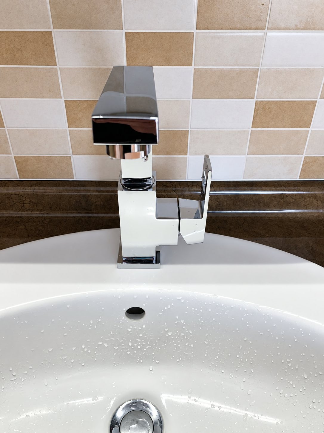 Robinet mitigeur chromé de style carré fixé au mur carrelé beige et marron, au-dessus d'une vasque ronde blanche avec des gouttes d'eau.