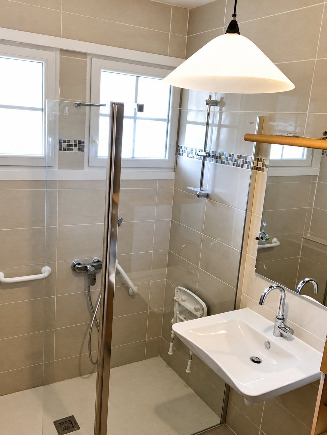 Salle de bain rénovée et adaptée aux personnes à mobilité réduite, avec paroi de douche en verre, siège rabattable, barres d'appui blanches, lavabo suspendu et suspension blanche.