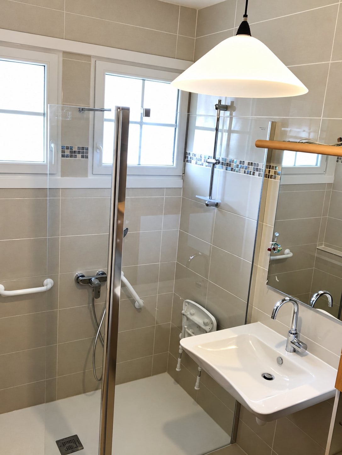 Salle de bain rénovée en carrelage beige avec douche à l'italienne vitrée, siège rabattable, barres d'appui blanches, lavabo suspendu et lustre blanc, photo prise en rotation.