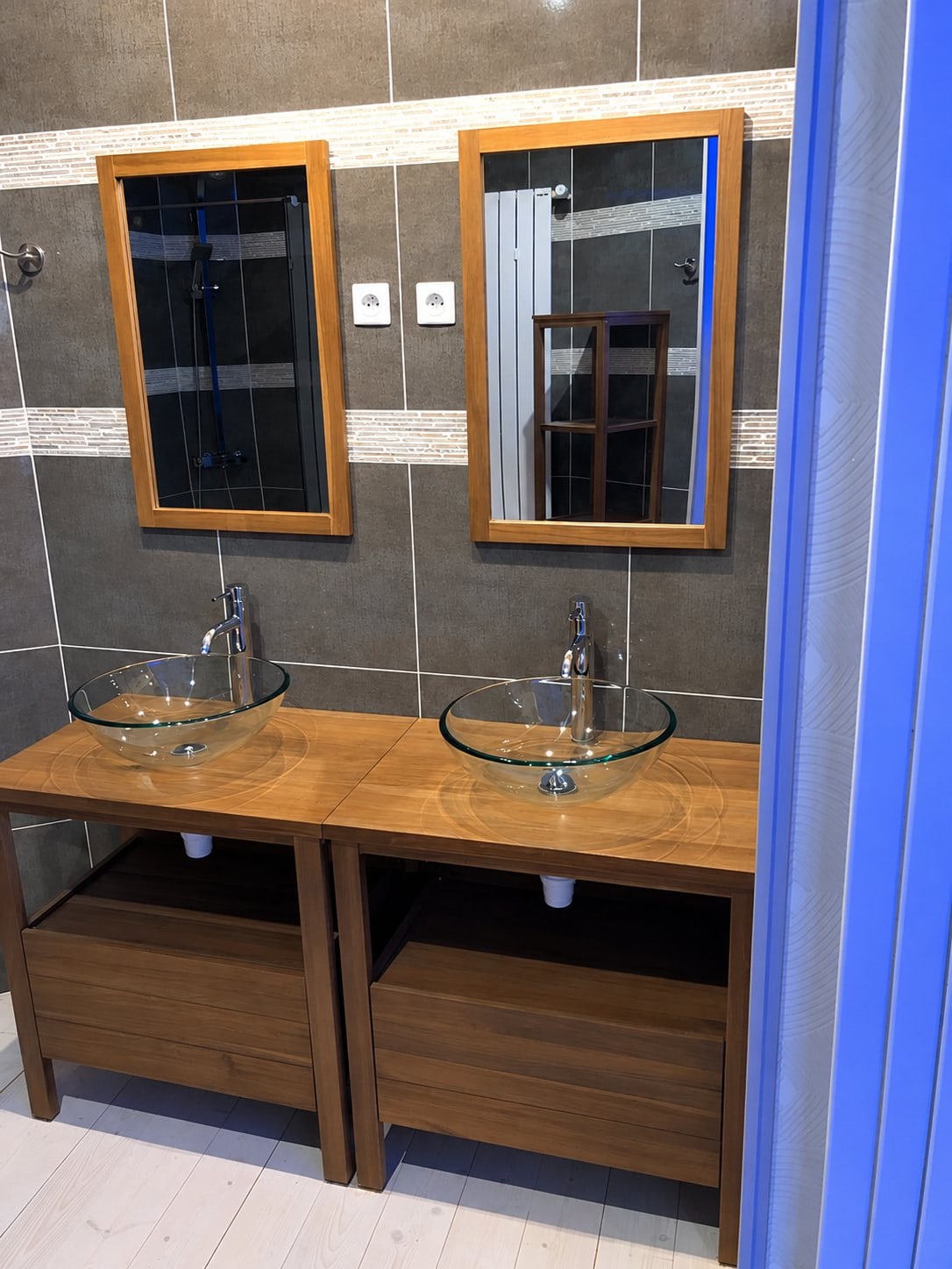 Salle de bain rénovée avec deux vasques en verre posées sur un plan en bois, deux miroirs à cadre bois et un mur en carrelage gris.