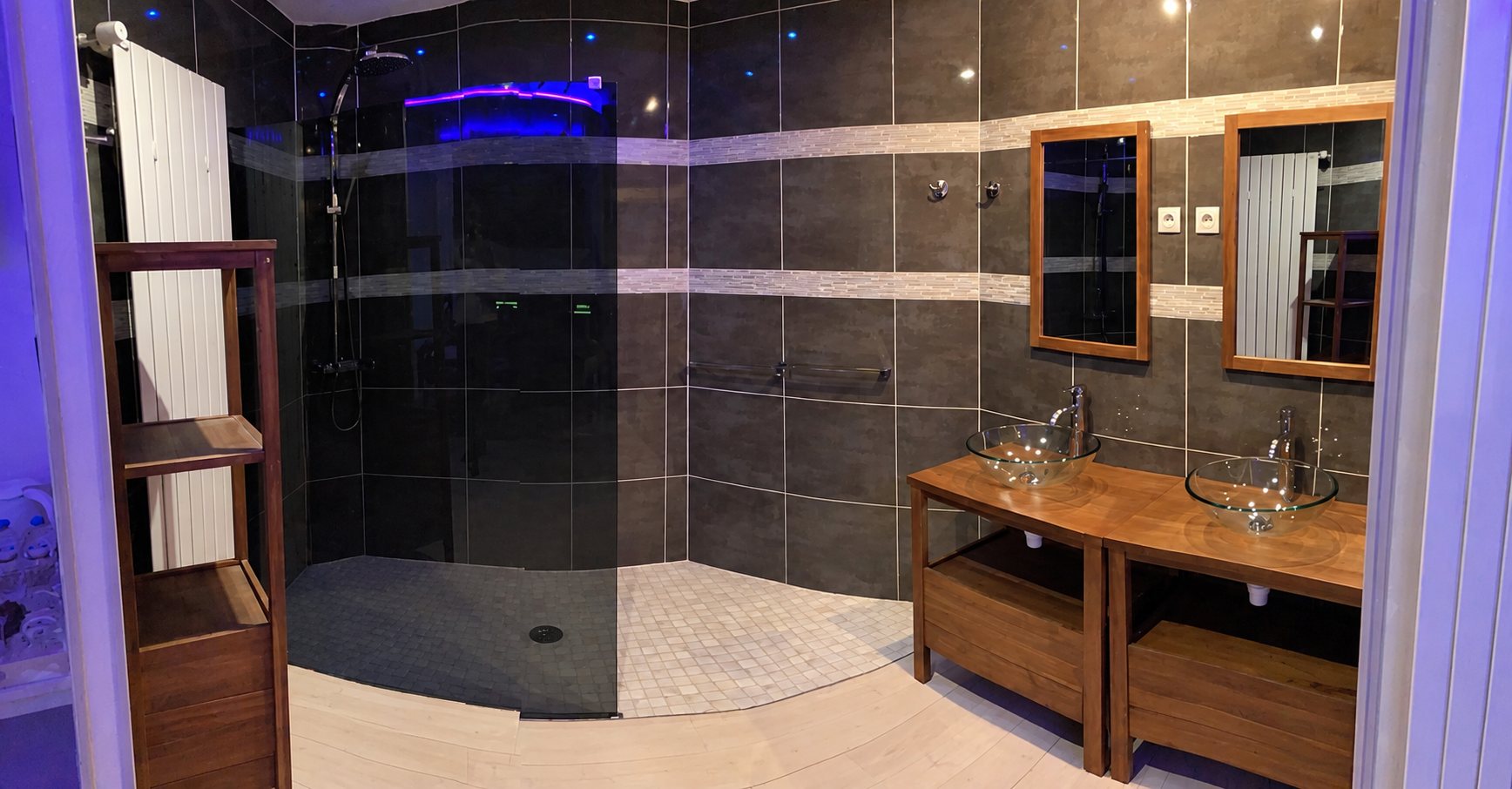 Salle de bain aux murs gris foncé avec grande douche à l'italienne, deux vasques en verre posées sur des meubles en bois et deux miroirs encadrés de bois.