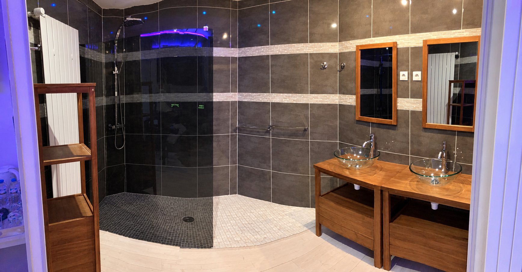 Salle de bain aux carreaux gris anthracite avec une grande douche à l'italienne, deux vasques en verre sur meubles en bois et miroirs encadrés de teck, éclairée par des lumières violettes.