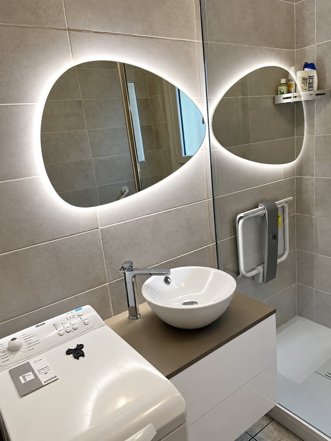 Salle de bain rénovée au carrelage gris avec un miroir ovale lumineux, une vasque blanche posée, un siège de douche rabattable et un lave-linge.