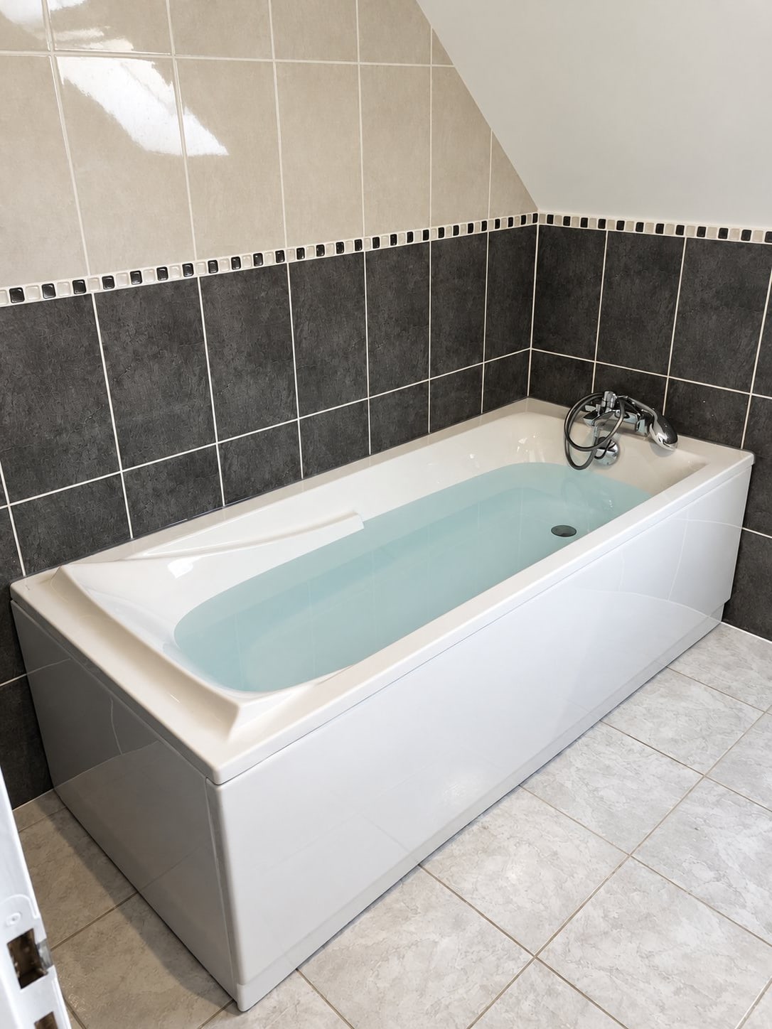 Baignoire rectangulaire blanche avec mitigeur chromé et douchette, entourée de carrelage gris foncé et beige avec une frise de mosaïque noire et blanche.