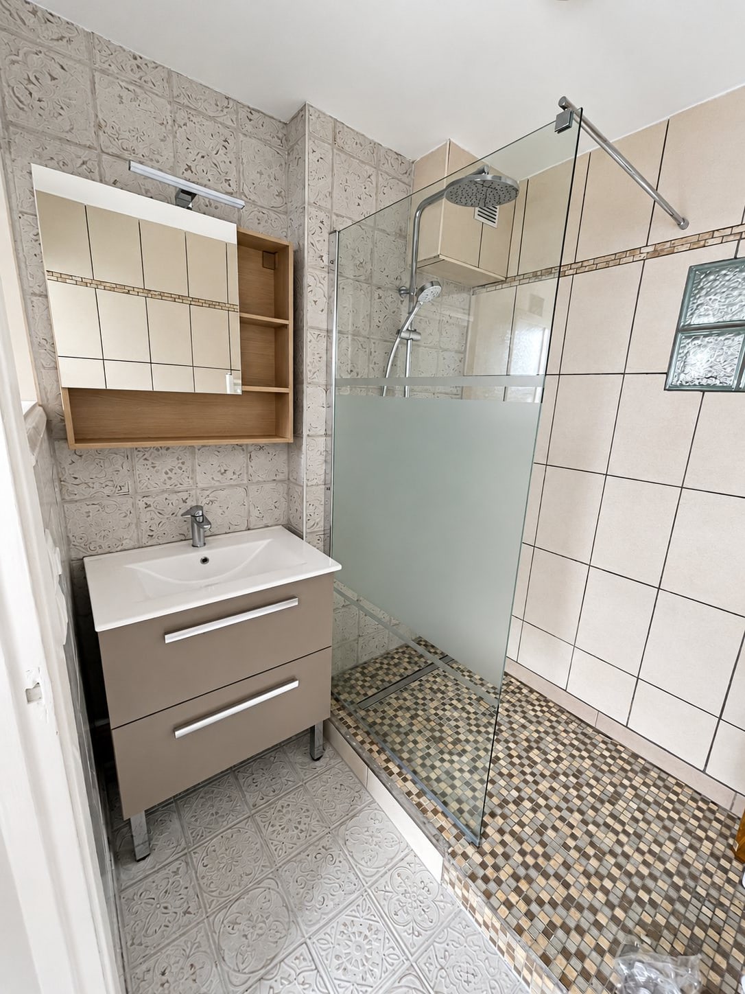 Salle de bain rénovée vue de dessus avec paroi de douche en verre dépoli, mosaïque murale, meuble vasque gris à deux tiroirs et miroir posé au sol.