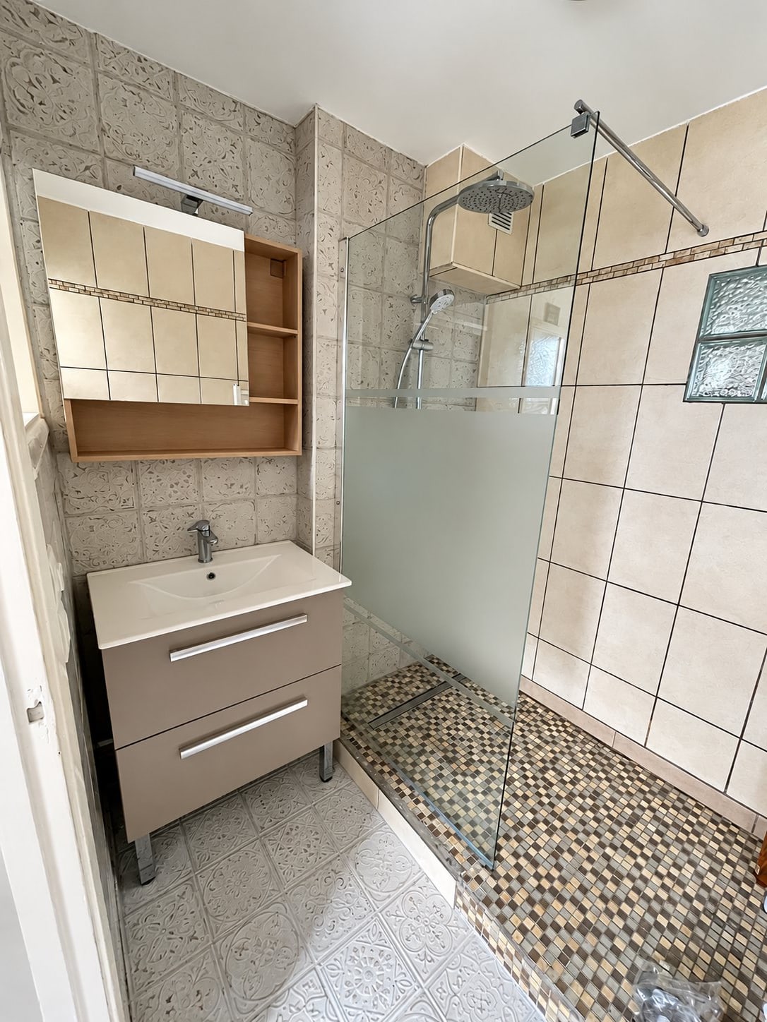 Salle de bain rénovée vue de côté, avec paroi de douche en verre dépoli, mosaïque murale, carrelage au sol et meuble vasque gris à deux tiroirs.