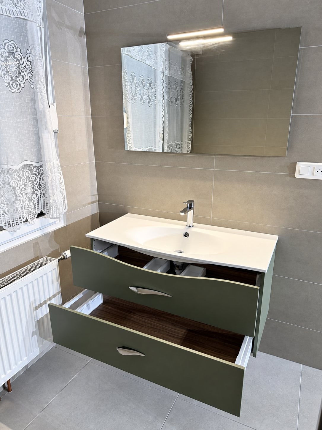 Salle de bain rénovée avec carrelage gris, miroir lumineux, meuble vasque vert à portes ouvertes, radiateur blanc et rideau de douche en dentelle.