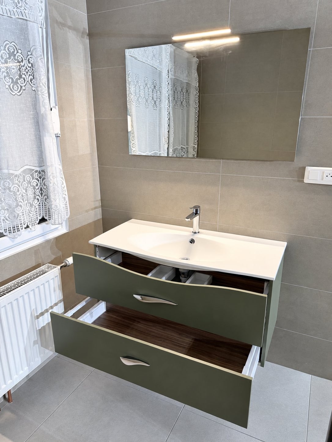 Salle de bain carrelée en gris avec un meuble vasque aux portes vertes ouvertes, un miroir rectangulaire, un radiateur blanc et un rideau de dentelle.