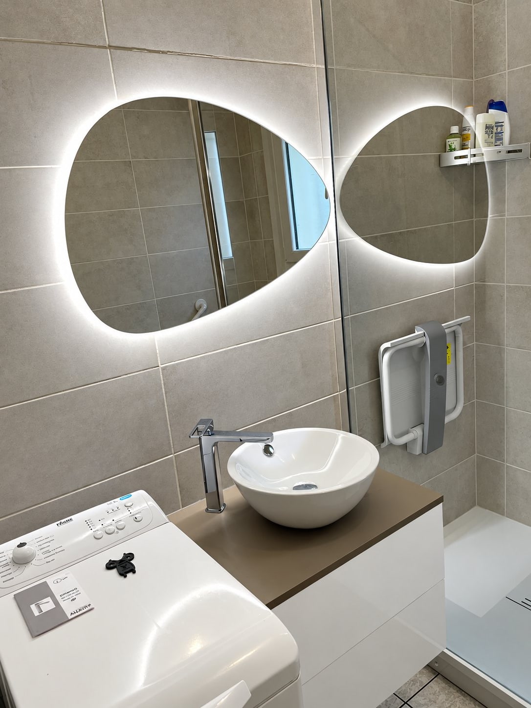 Vue plongeante d'une salle de bain rénovée avec carrelage gris, miroir organique rétroéclairé, vasque blanche posée et siège de douche rabattable.