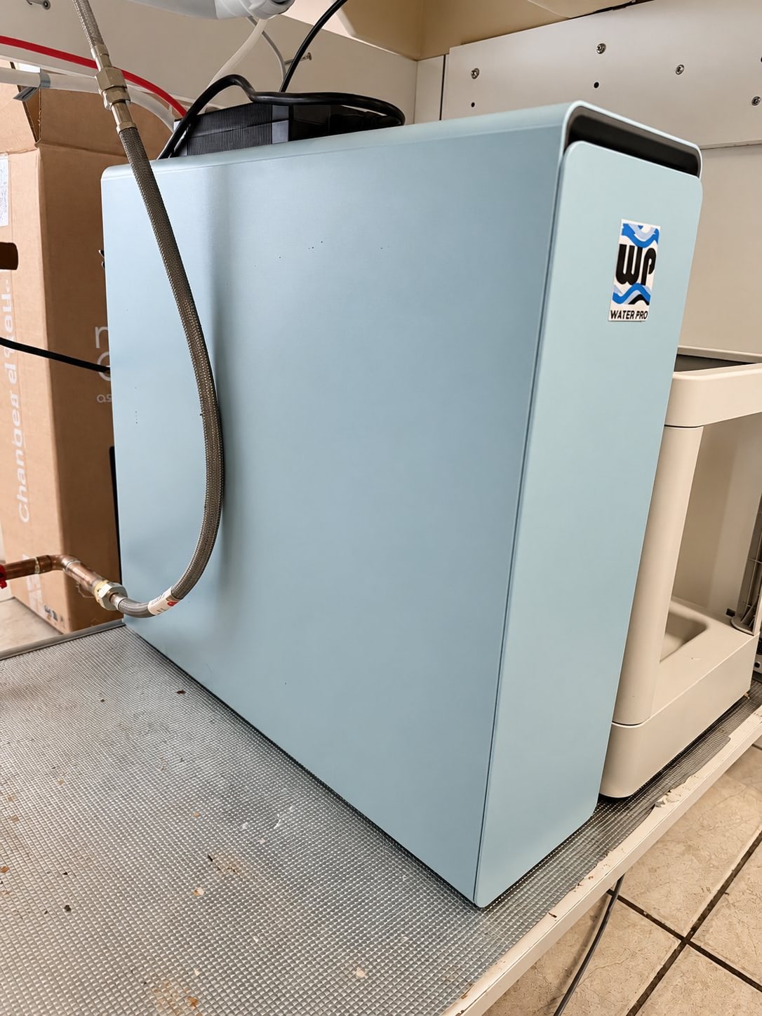 Un boîtier de filtration d'eau bleu pâle, portant un autocollant Water Pro, est raccordé par un flexible tressé et posé près d'un carton et d'un sol carrelé.