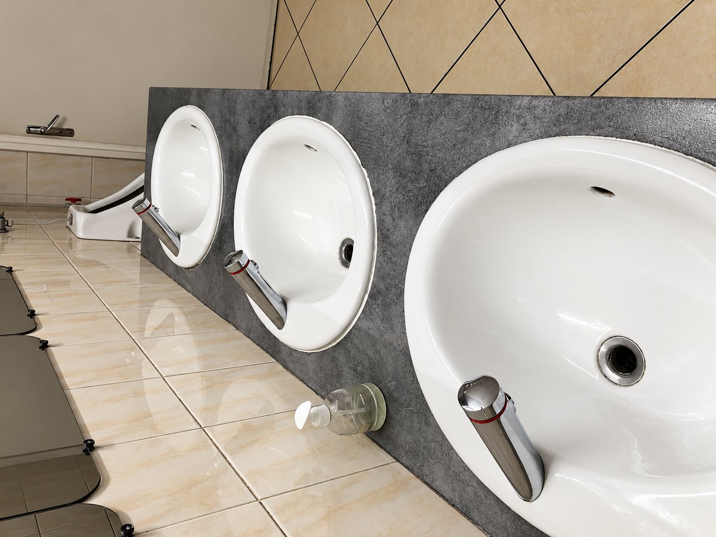 Trois lavabos ovales blancs avec robinets chromés sont encastrés dans un plan gris effet béton, dans des sanitaires au sol carrelé beige.