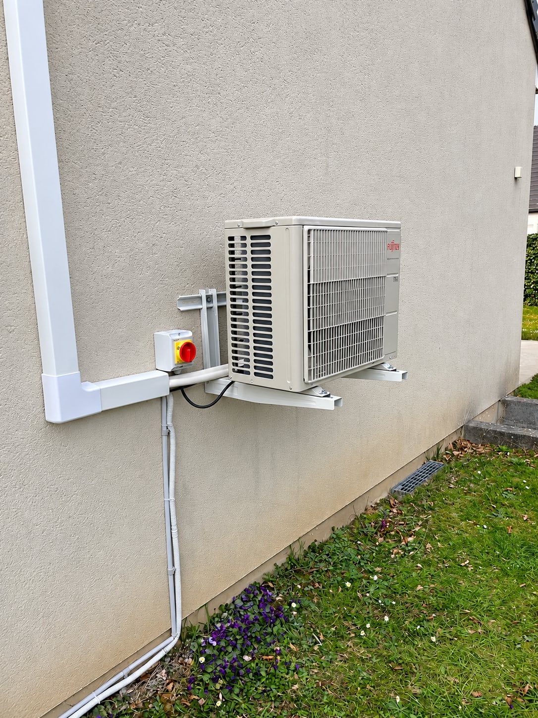Une unité extérieure de climatisation Fujitsu fixée sur supports au sol d'une terrasse, avec interrupteur de sécurité rouge et goulottes blanches, à côté d'une pelouse.