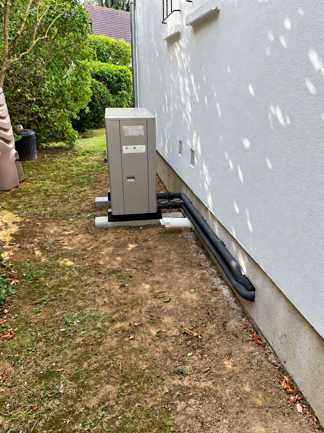 Une unité extérieure de pompe à chaleur grise est posée sur des plots contre le mur d'une maison, reliée par des tuyaux isolés noirs.