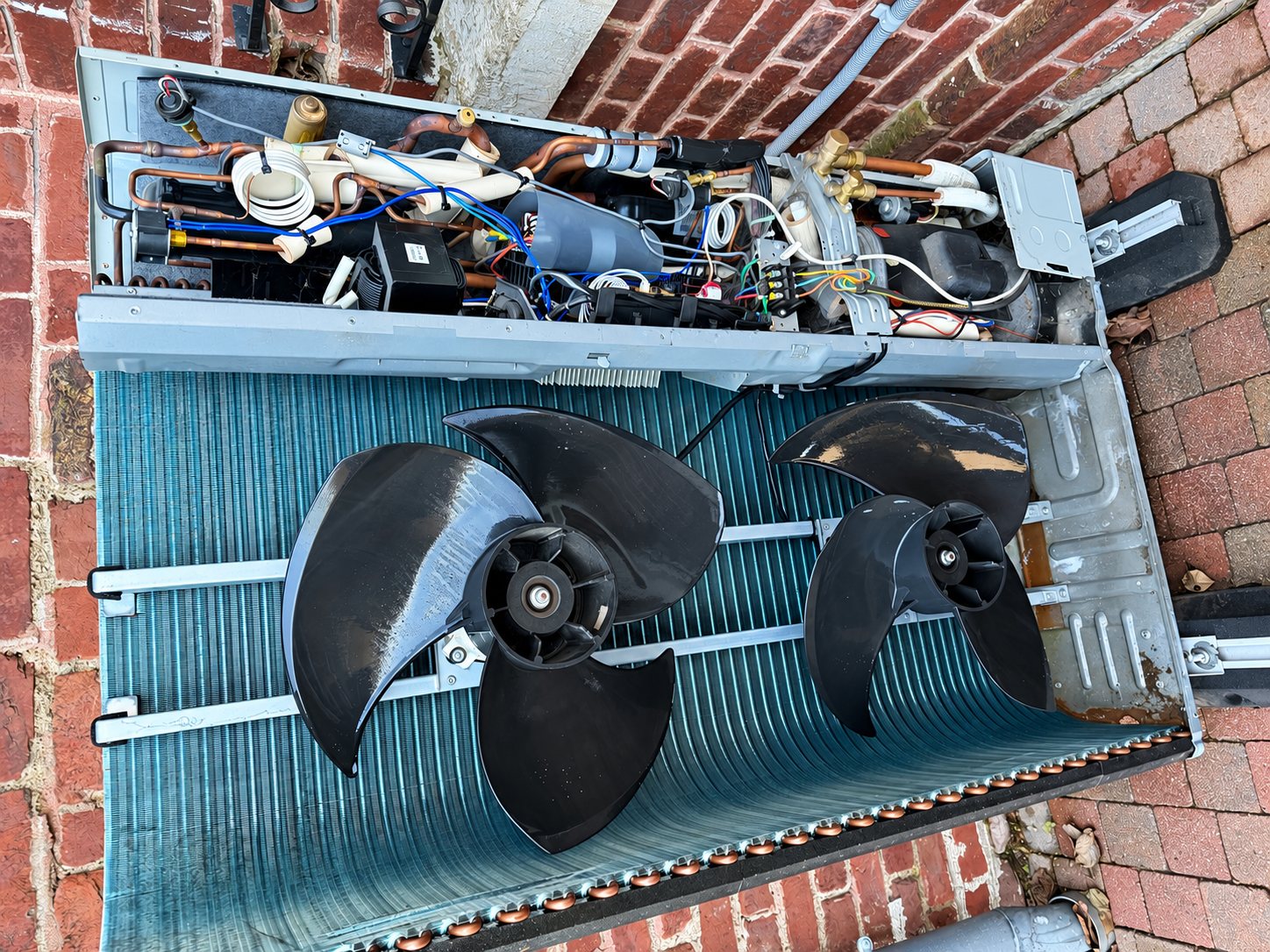 Vue de dessus d'une unité extérieure de pompe à chaleur démontée posée sur un sol en briques, laissant voir deux ventilateurs noirs, l'échangeur et les tuyaux en cuivre.